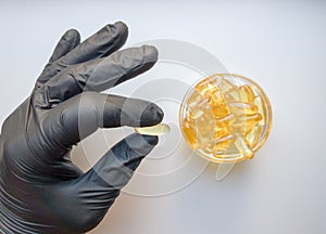 Fish oil capsules in hand in a black medical glove on a white background, the hand takes one tablet from a saucer or plate. Hand