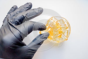 Fish oil capsules in hand in a black medical glove on a white background, the hand takes one tablet from a saucer or plate. Hand