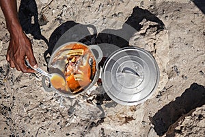 Fish soup or stew cooked from freshly catch seafood in aluminium pot, closeup detail to local man hand from above, sun