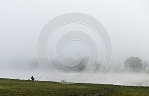 Fisher man fishing on a river bank at misty foggy sunrise
