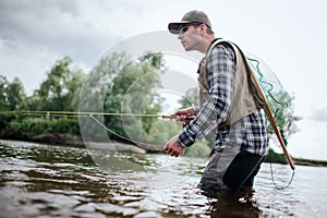Fisherman in action. He stands in water and holds fly rod in one hand and spoon in the other one. Also adult has a