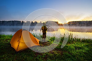 Fisherman on the bank of foggy river near an orange tent in the early morning