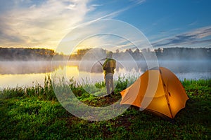 Fisherman on bank of foggy river near an orange tent in the early morning