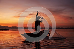 Fisherman casting his net on during sunset. Silhouette Asian fisherman on wooden boat casting a net for freshwater fish.