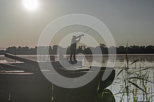 Fisherman fishing in early serene morning. Man black silhouette resting with rod on river or lake with sun light
