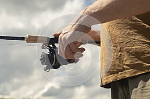 Fisherman hands holding spinning reel rod or spoon bait and fishing close up. Summer sky with clouds on background