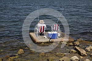 Fisherman in a hat sitting on a chair with a red and white stripe is fishing with a fishing rod on the sea or lake