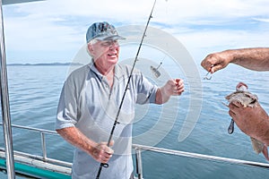 Fisherman helping caucasian tourist on fishing charter boat taking hook out of snapper fish - at Doubtless Bay, Far