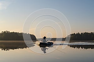 Fisherman in an inflatable boat rowing on a calm forest lake, at dawn, against a background of sky and trees. Beautiful