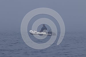 Fisherman in inflatable fishing boat with outboard motor moving in the dense fog.