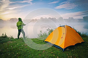Fisherman by lake with orange tent on foggy morning