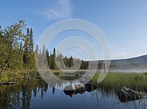 Fisherman man catching fish at lake Sjabatjakjaure in Beautiful sunny morning haze mist in Sweden Lapland nature. Mountains, birch