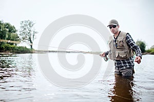 Fisherman in protection clothes is stnading in water and fishing. He holds fish rod in one hand and pulls spoon with