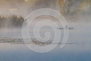 Fisherman in a punt boat fishing with rods, thick fog covers still surface of a river, bulrush, cane and willows grow on a bank