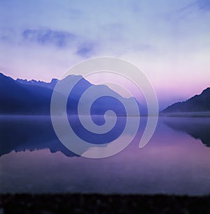 A fisherman rowing among the fog on the lake of Silvaplana in the Engadin valley at sunrise with mountains reflecting in the water