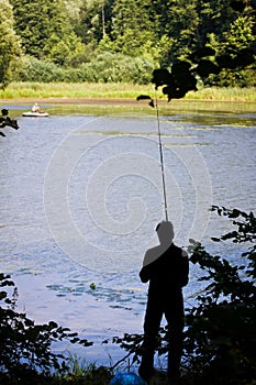Fisherman`s figure with  a fishing rod on a bank of little natural lake in forest, calm breeze on water surface