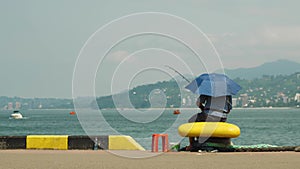 A fisherman sits on the city waterfront and fishes in the sea. A man with a fishing rod is hiding from the sun under an
