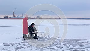 Fisherman sits on frozen river and cathes fish with help winter fishing rod and next to pretty young woman watches for fisherman