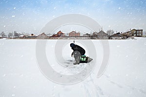A fisherman sits near an ice hole on a frozen snow-covered river in a snowfall