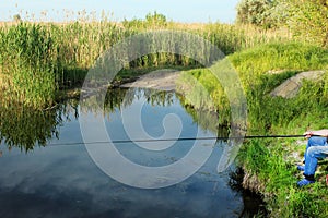 A fisherman sits on the shore of a lake with a fishing rod in his hands against the backdrop of wild nature on a bright sunny summ