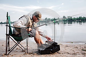 Fisherman is sitting in folding chair and leaning forward to opened black plastic box. Guy holds fly rod and plastic