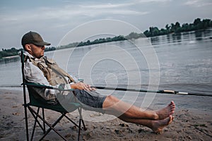 Fisherman is sitting in folding chair and sleeping. His cap is lying on eyes. Man holds fly rod in hands. He is fishing