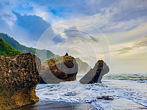 A fisherman sitting on a rock formation while big long waves flo