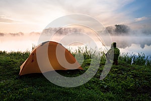 Fisherman sitting on shore of foggy lake near an orange tent