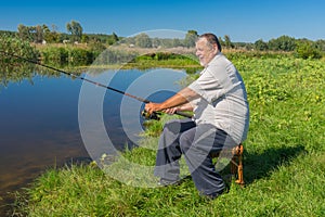 Fisherman sitting on a wicker stool with spinning rod and ready to catch fish in small river Merla in central Ukraine