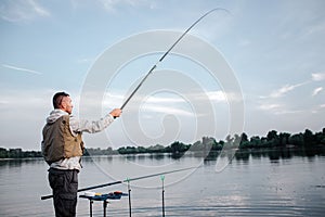 Fisherman is standing near watr and holding fly rod with right hand. He has it up really high. Man looks straight