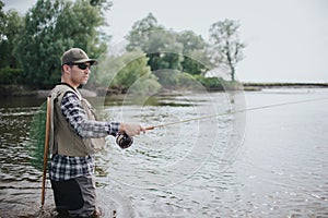 Fisherman in sunglasses is standing in water. He looks to the right. Guy has fishing net on the back. Man is holding fly