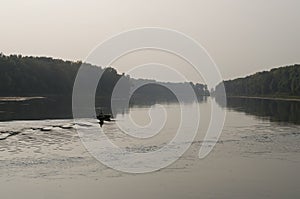 Fisherman are swiming on the boat. Wide river flowing across green forest. Fall. Evening. Reflections of trees in the calm water.