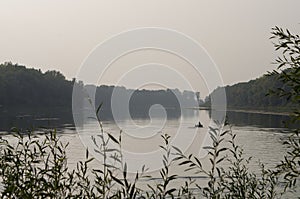 Fisherman are swiming on the boat. Wide river flowing across green forest. Fall. Evening. Reflections of trees in the calm water.