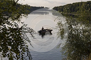Fisherman are swiming on the boat. Wide river flowing across green forest. Fall. Evening. Reflections of trees in the calm water.