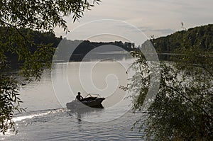 Fisherman are swiming on the boat. Wide river flowing across green forest. Fall. Evening. Reflections of trees in the calm water.