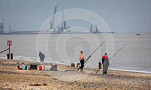 Fishermen fishing off the beach with cranes in Yarmouth Harbour in the back ground, near Lowestoft, Norfolk, UK