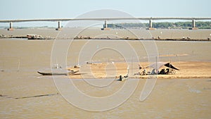 Fishers are sitting under a tarpaulin on the river sandbank ; Motorbikes and bicycles crossing bamboo bridge over the Mekong river