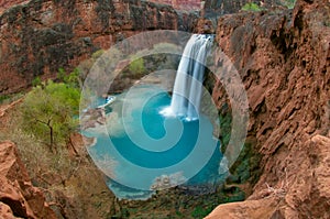 A bird`s eye view of the 100ft Havasu Falls and its beautiful deep blue pool beneath, surrounded by thick, green vegetation.