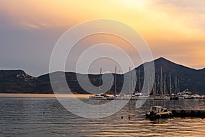 Fishing and sailing boats docked in Adamas Port on Milos Island, Greece during golden hour sunset, with calm flat sea