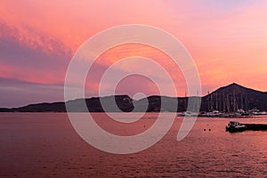 Fishing and sailing boats docked in Adamas Port on Milos Island, Greece during vivid pink sunset, with calm flat sea