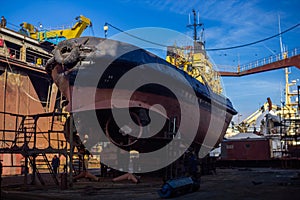 Fishing vessel maintenance in shipyard drydock. Workers repair, repaint hull, propeller, docked boat under clear sky