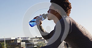 Fit african american man exercising in city, resting on footbridge, drinking from water bottle