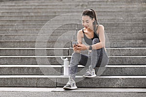 Fit asian girl using smartphone while sitting on stairs after training outdoors
