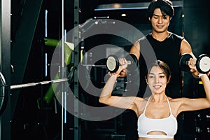 Fit Asian woman lifting dumbbells with a smile, supported by her personal trainer