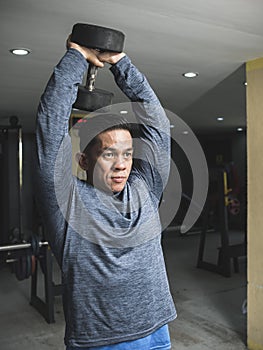 A fit athletic asian guy in a sweatshirt does standing overhead dumbbell tricep extension with both arms. Starting position.