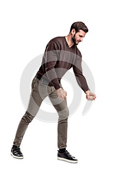 A fit bearded young man in casual clothes tries to pull an invisible rope with all his strength from low grip.