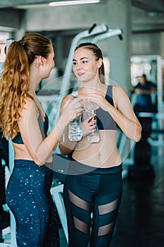 Fit beautiful young woman athletes in fitness clothes relaxing after workout in gym. Couple women standing holding water in bottle