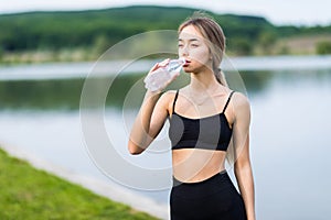 Fit beautiful young woman drinking water after workout by the lake. Teenage runner at the beach with bottle of water refreshing