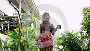 Fit biethnic woman lifts dumbbells in upscale outdoor gym, surrounded by rich tropical green plants. Athletic female