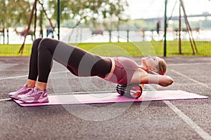 Fit blond woman training on mat outdoor summer day, using foam roller massager on her shoulders for relaxation